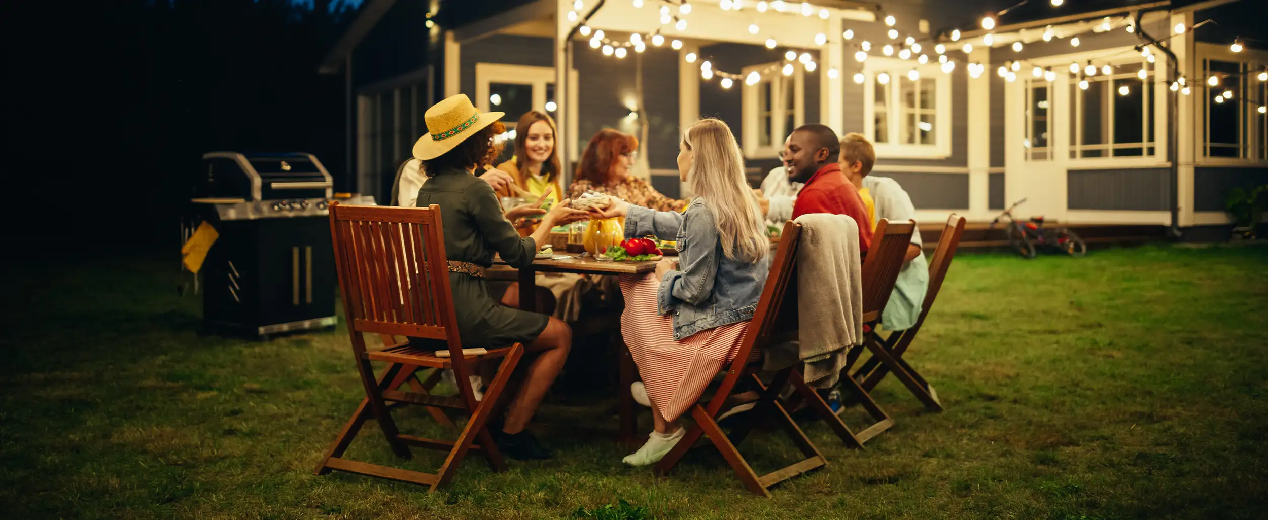 Family and Multicultural Friends Celebrating Outside at Home in the Evening. Group of Children, Adults and Old People Gathered at a Table, Having Fun Conversations. Eating Barbecue and Vegetables.